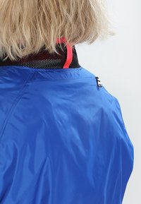 Modrá voděodolná bunda s hladkou texturou, vybavená černým síťovaným límcem a růžovým akcentem. Obsahuje malé zipové detail na zadní straně.