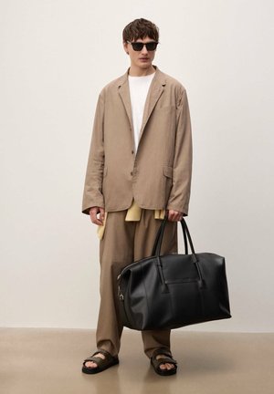 Ung mand iført beige jakkesæt, hvid skjorte, sorte solbriller og sandaler, der holder en stor sort læderduffeltaske, stående foran en ensfarvet baggrund.