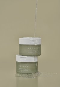 Groene ronde potten met witte deksels, gelabeld "NEEDLY Cicachid Koelpad." Water spettert naar beneden en benadrukt de textuur en helderheid van de containers.