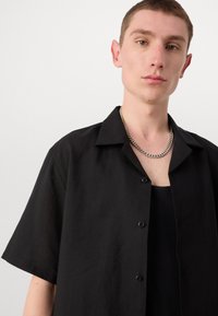 Camisa negra de manga corta con un ajuste relajado, hecha de tela ligera. Presenta un cierre de botones en la parte delantera y un cuello, con una apariencia texturizada.