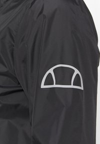 Svart jacka med en slät, glänsande yta; har en stor, grå böjd logotyp på vänster arm; lättviktig, minimalistisk design.