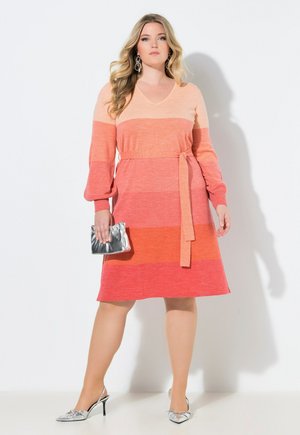 Nainen pitkähihainen, väriblokattu vaaleanpunaisesta koralliin oleva mekko, jossa on vyö, pitää hopeista käsilaukkua ja käyttää hopeisia korkokenkiä ja korvakoruja.