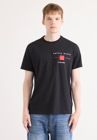 Zwarte katoenen t-shirt met een rood-witte grafische print. Ronde hals, korte mouwen en een losse pasvorm. Gepaarde met blauwe denim jeans.