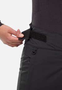 Hand, die einen schwarzen verstellbaren Seitenriemen mit Klettverschluss an der Taille eines schwarzen Kleidungsstücks über einem dunklen Langarmshirt anpasst.