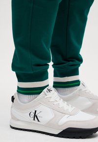 Fehér és szürke sportcipők fekete talppal, Calvin Klein felirattal, zöld jogger nadrággal párosítva, fehér és zöld csíkos szegéllyel.