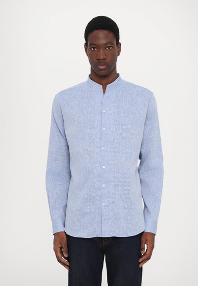 Camicia azzurro chiaro a maniche lunghe con colletto alla cinese, caratterizzata da una texture delicata e bottoni bianchi sul davanti. Indossata con jeans scuri.