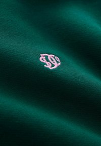 Zielona tkanina o gładkiej fakturze z małym różowym haftowanym logo w dolnym rogu. Zbliżenie ukazuje szczegóły materiału.