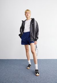 Svart jacka med dragkedja och vita ränder, vit t-shirt, blå shorts och svarta skor med ljusbruna sulor. Står på en blå matta.