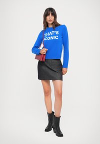Blå strikkegenser med hvit tekst "THAT'S ICONIC," svart denimskjørt og svarte hælboots. Rød crossover-veske gir et fargeakkcent.
