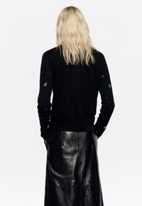 Melnā džemperī ar garām piedurknēm, kas dekorēts ar rotājumiem uz rokām, apvienots ar garu melnu ādas svārku.