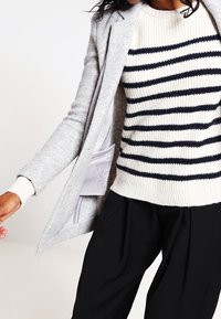 Blazer de mezcla de lana gris sobre un suéter de punto crema con rayas marinas. Pantalones negros holgados completan el look.