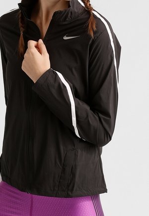 Person trägt eine schwarze Nike-Jacke mit weißen Streifen an den Ärmeln und violette strukturierte Leggings, hält den Jackenkragen mit einer Hand.