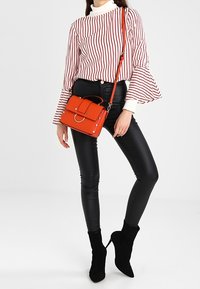 Sac à main structuré rouge avec une poignée supérieure et une sangle ajustable, porté par une personne portant une blouse rayée et un legging en simili cuir noir.