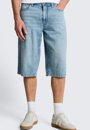 Mann trägt hellblaue Jeans-Knielange Shorts, weiße Socken und cremeweiße Sneakers, steht mit einer Hand in der Tasche.
