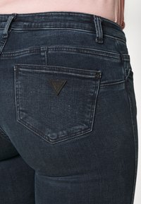 Jeans de mezclilla de un azul oscuro con un bolsillo trasero que presenta un pequeño parche en forma de triángulo negro, llevados con una blusa de color rosa claro.