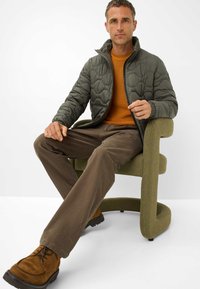 Gequiltde olijf groene jas, oranje sweater, bruine broeken en tan suède schoenen gedragen terwijl hij/zij op een olijf groene, gebogen stoel zit.