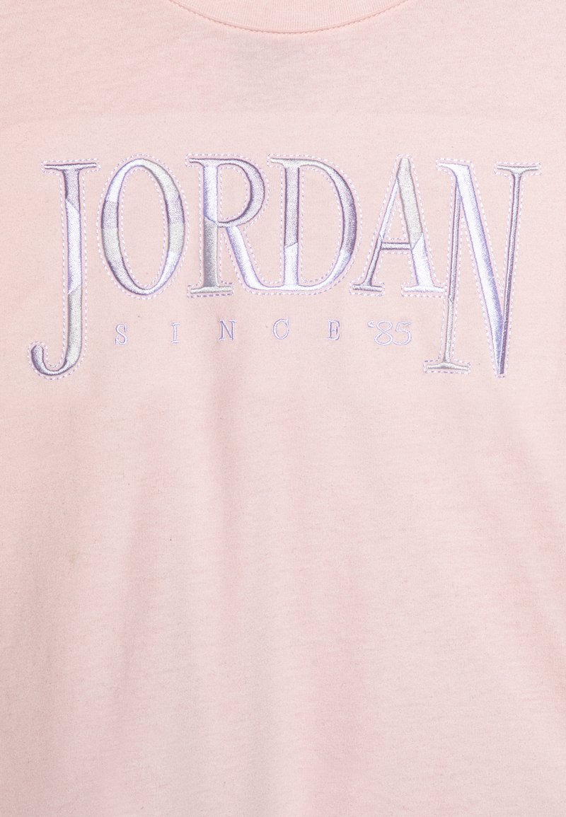 T-shirt di cotone rosa con la scritta "JORDAN" in ricamo testurizzato blu chiaro con un motivo sottile, e "SINCE '85" in testo più piccolo sotto.