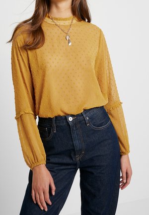 Kvinde iført en sennepsgul tekstureret langærmet bluse, stoppet ned i mørkeblå højtaljede jeans, med en sølvvedhæng halskæde.