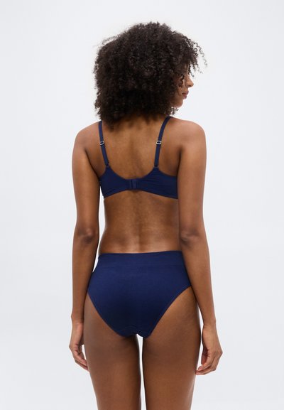 Γυναίκα με σγουρά μαλλιά που φοράει μπλε σκουρο μπρά και ταιριαστό εσώρουχο με ψηλή μέση, σε εικόνα από πίσω.