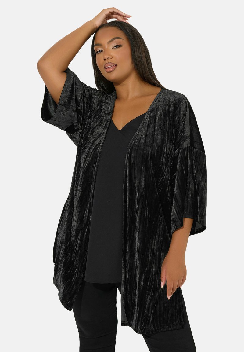 Zwarte, gestructureerde fluwelen kimono met brede mouwen en een open voorkant, gedragen over een zwarte top. Heeft een ontspannen pasvorm en valt elegant.