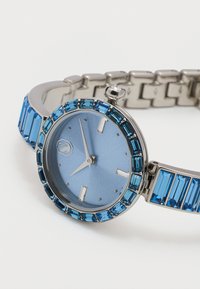 Ασημένιο ρολόι χειρός με ανοιχτό μπλε καντράν, ασημένιους δείκτες και μπλε ορθογώνιες πολύτιμες πέτρες γύρω από το στεφάνι και τους κρίκους του βραχιολιού σε λευκό φόντο.