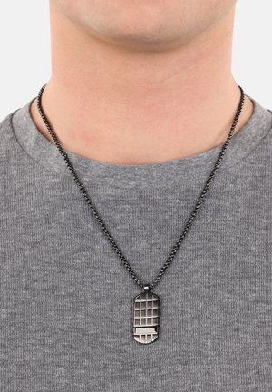 Collana con medaglietta in metallo con un motivo a griglia, appesa a una catena nera, indossata sopra una maglietta grigia con texture.