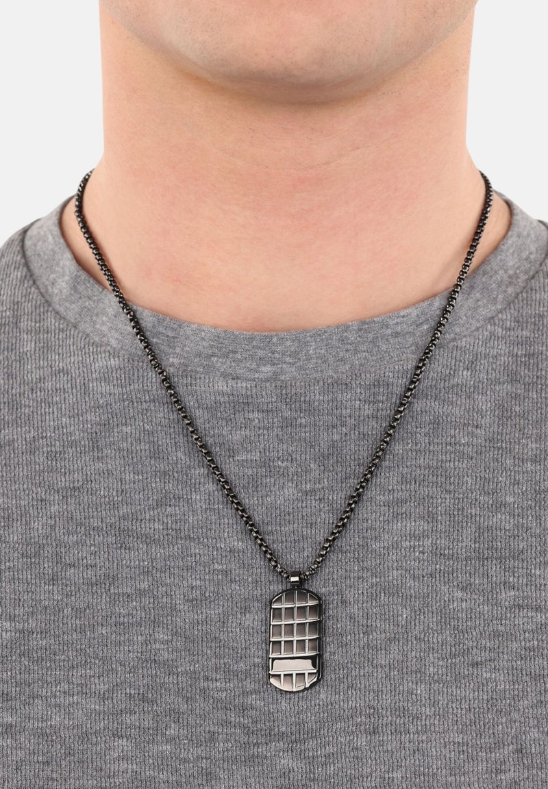 Collana con medaglietta in metallo con un motivo a griglia, appesa a una catena nera, indossata sopra una maglietta grigia con texture.
