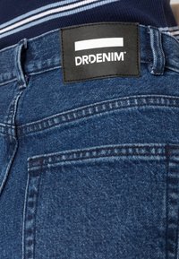 Jeans de ganga azuis com um acabamento texturizado, apresentando uma etiqueta de couro com o logótipo "DRDENIM", costuras visíveis e um design padrão de bolso traseiro.