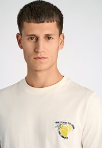 T-shirt corto a maniche corte color crema con grafica di un limone e testo ricamato sul lato sinistro del petto. Materiale in cotone morbido con scollatura a girocollo.