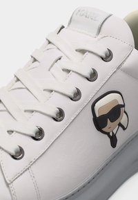 Λευκό δερμάτινο αθλητικό παπούτσι με λευκά κορδόνια, μεταλλικά κουμπώματα και έμβλημα καρτούν με χαρακτήρα που φοράει μαύρα γυαλιά ηλίου στο πλάι.