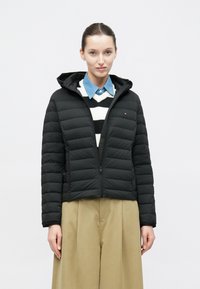 Fekete, bélelt dzseki kapucnival, elöl cipzárral, bordázott mandzsettával és kis logó részlettel. Csíkos pulóver és gallér fölött viselve.