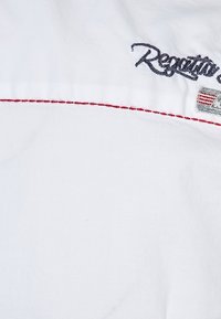 Biały materiał z czerwonymi detalami szwów. Zawiera haftowany napis "Regatta" w granatowym kolorze oraz małą teksturowaną metkę z motywem flagi.
