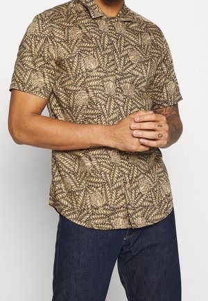 Uomo che indossa una camicia a maniche corte con bottoni con motivo di foglie e tigri beige e marrone, abbinata a jeans blu scuro.