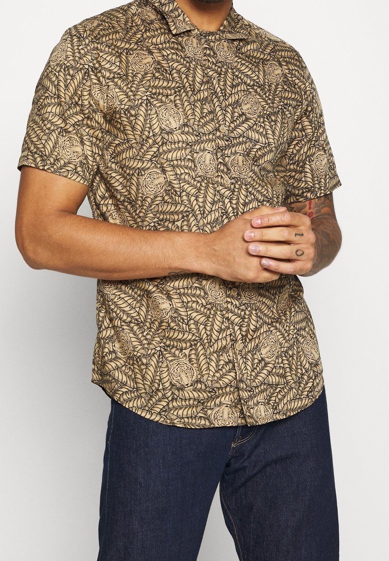 Homme portant une chemise boutonnée à manches courtes avec un motif feuilles et tigres beige et marron, associée à un jean bleu foncé.