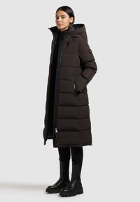 Manteau doudoune long, brun foncé, avec une capuche, doté d'une fermeture éclair sur le devant, de poches latérales et d'un design matelassé. Assorti à des bottes noires et des leggings.