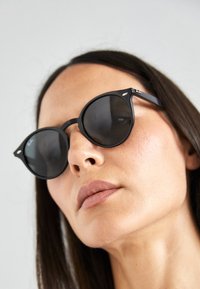 Ochelari de soare negri cu lentile rotunde, cadru mat, accente metalice, design minimalist și siluetă elegantă, pe un fundal neutru.