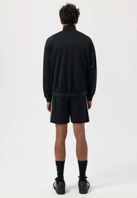 Svart atletisk jacka med hög krage, kombinerad med matchande shorts med sidostraper. Bärs med svarta strumpor och skor.