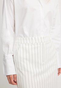 Witte blouse met lange mouwen, gecombineerd met een witte rok met dunne verticale zwarte strepen en een gladde textuur.