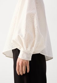 Blusa bianca a maniche lunghe con motivo ad occhio di pernice ricamato, maniche voluminose e polsini con bottoni. Indossata su stoffa nera.