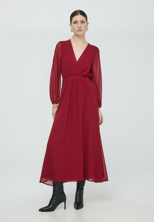 Lungo vestito bordeaux con scollo a V, stile avvolgente e maniche lunghe in tessuto trasparente. Abbinato a stivali neri con punta a silo. Texture liscia, design fluido.