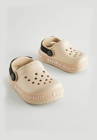 Zuecos de goma beige con agujeros de ventilación, correa negra en el talón y branding blanco "BAKER by TED BAKER" en el lateral. Forro suave visible en el interior.