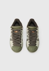 Zaļas un bēšas sporta kurpes ar apaļu gumijas pirksta daļu, biezām brūnām šņorēm un smalku zīmola apzīmējumu sānos un mēlē.