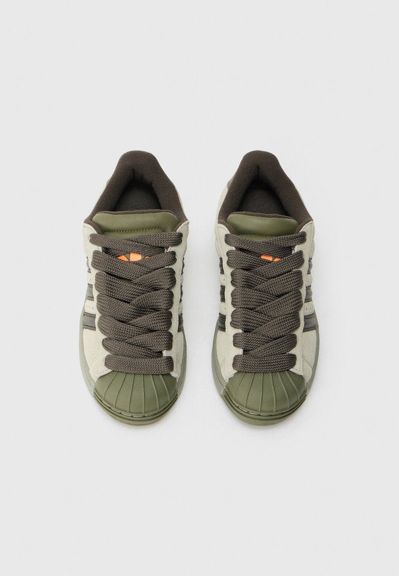 Baskets vertes et beiges avec un embout en caoutchouc arrondi, des lacets bruns épais, et un marquage subtil sur le côté et la languette.
