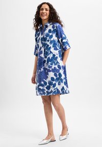 Blue and white floral linen dress with short sleeves, button front, and side pockets. Model wears silver slip-on shoes.