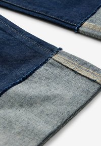 Jeans in denim con parte superiore blu scuro e polsini blu chiaro, con bordi sfrangiati e cuciture a contrasto. Tessuto testurizzato con trama visibile.