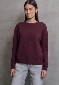 Pull en tricot bordeaux avec manches longues, col rond, poignets et ourlet côtelés. Associé à un jean gris clair. Texture lisse, design minimaliste.