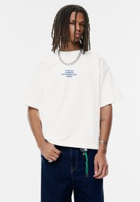 Weißes, übergroßes T-Shirt mit blauer Textdruck, kombiniert mit dunklen Jeans und einer silbernen Kettenhalskette, was einen lässigen Schnitt und ein minimales Design zeigt.