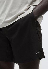 Μαύρο αθλητικό σορτς από λείο, ελαφρύ ύφασμα, με μικρό λευκό λογότυπο. Ο σχεδιασμός είναι μίνιμαλ με τσέπες στην πλαϊνή ραφή.