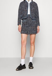 Completo giacca e gonna in tweed di colore navy, con fili multicolori, due tasche frontali e bordi blu a contrasto. Abbinato a scarpe nere lucide.