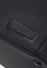 Sac en cuir noir texturé avec une étiquette rectangulaire noire distinctive portant un logo en relief. Surface lisse avec des détails de surpiqûres.